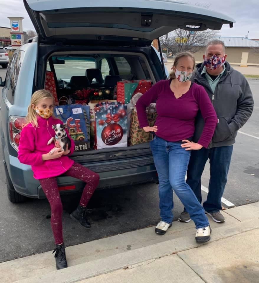 Man, woman, young girl, and their puppy all in masks, in front of a full trunk of gift bags for drop off